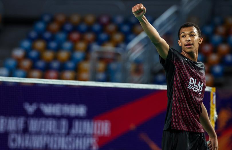 Юный игрок в тёмной форме вскинул кулак у сетки, на баннере — надпись BWF World Junior Championships (фото: BWF/Badmintonphoto — Raphael Sachetat)