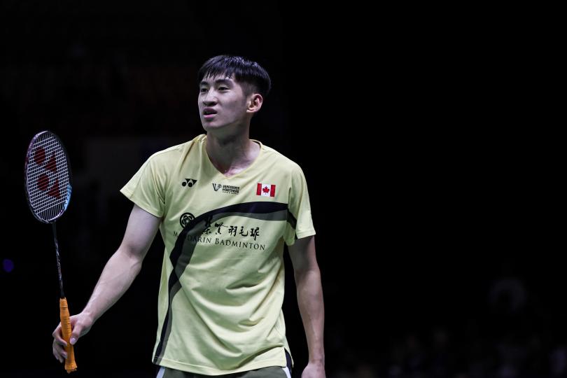 Виктор Лай в жёлтой майке с флагом Канады и надписью Mandarin Badminton держит ракетку с оранжевой рукояткой на тёмном фоне, кадр чемпионата мира (фото: BWF/Badmintonphoto — Jnanesh Salian)