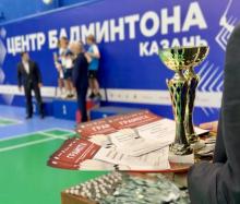 Кубок и стопка красных грамот на столе, на заднем плане — пьедестал почёта и награждение у стены «Центр бадминтона. Казань». Фото: НФБР (nfbr.ru).